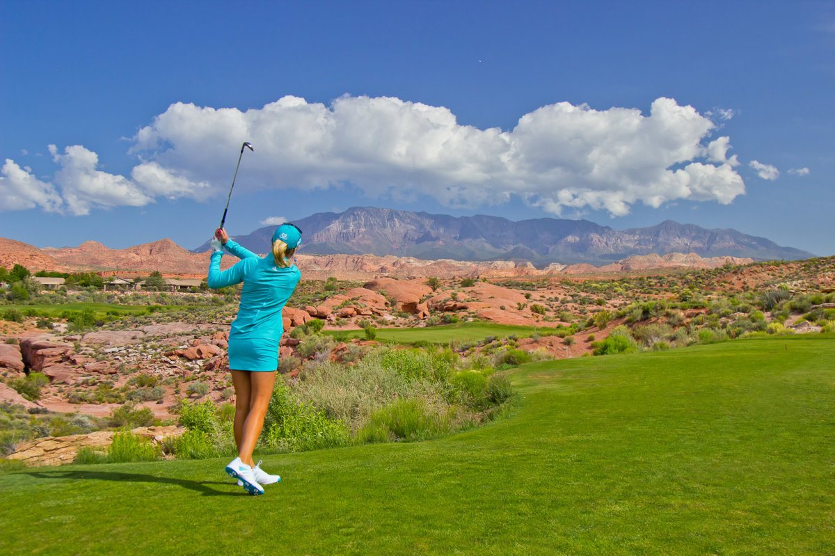 Female golfer midswing under blue sky with fluffy clouds.