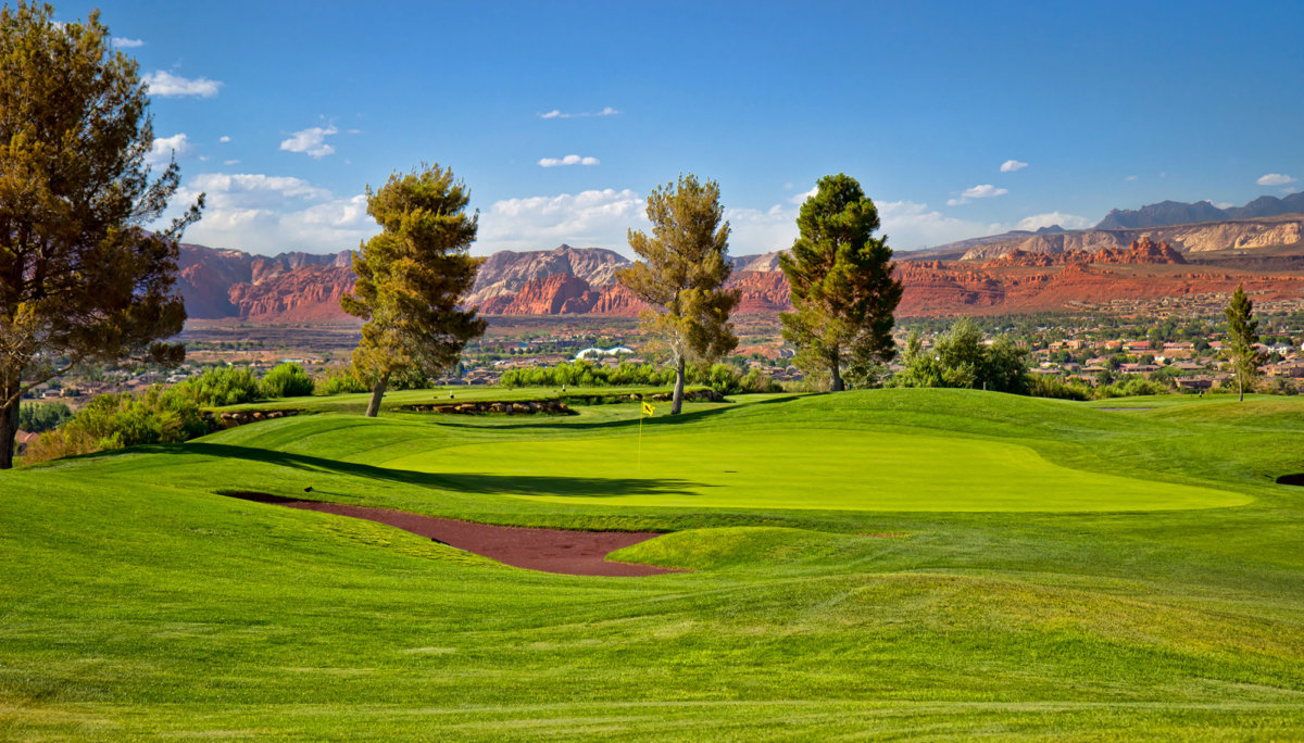 Green fairway with pine trees and red rock formation in background.